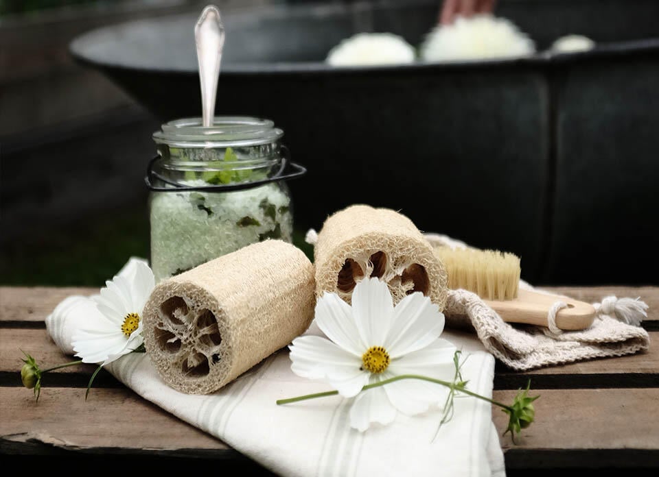 En person bakom en burk med hemlagad sockerpeeling, blommor och en luffa.